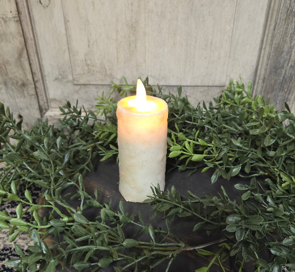 A realistic moving flame votive candle with an ivory color, placed on a dark container, surrounded by greenery. The candle is lit, and the flame is visible, creating a warm and inviting atmosphere.