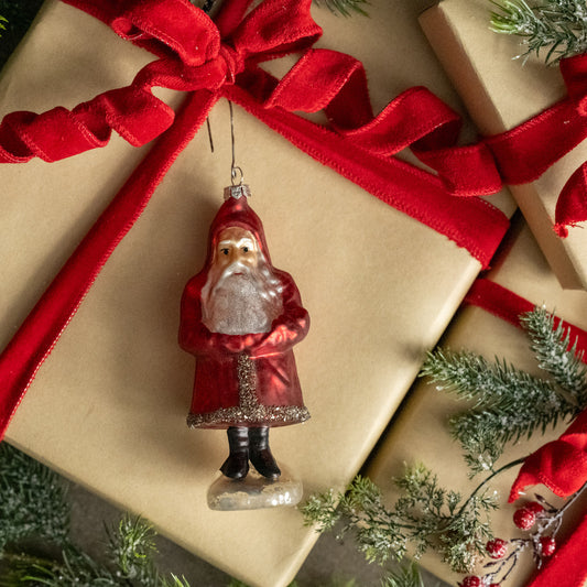 Christmas ornament of a Santa Claus figure on a gift box with red ribbon and greenery.