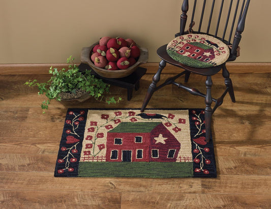 A hooked rug with a red house design, placed on a wooden floor next to a wooden rocking chair and a bowl of fruit.