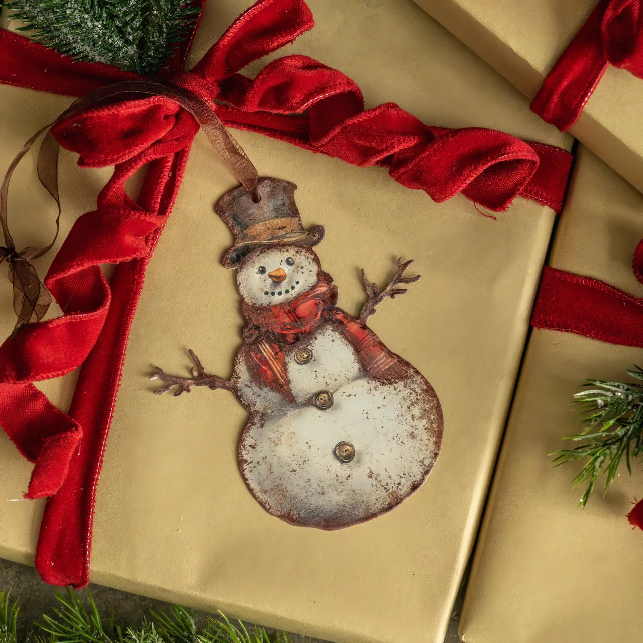 A decorative snowman ornament with a top hat and scarf, attached to a kraft gift box tied with a red velvet ribbon. Other wrapped boxes and evergreen branches are partially visible around it.
