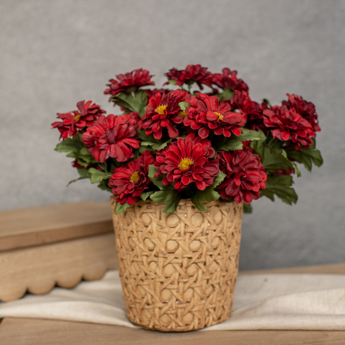 An artificial floral arrangement in a woven basket featuring vibrant red zinnia flowers and green leaves.