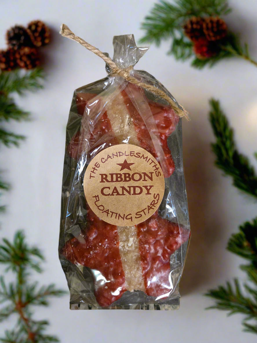 Packaged ribbon candy floating candles with a label on a wooden surface surrounded by sprigs of greenery.