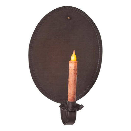 A black metal candle sconce with a lit candle inside, mounted on a circular black base with a primitive finish.