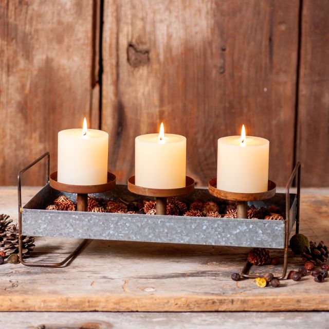 Three pillar candles in a black metal 3-pillar candle holder tray with a rustic appeal, placed on a wooden surface with pinecones and decorative branches around.