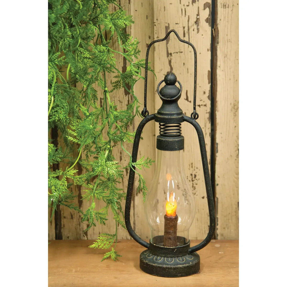 A rustic black metal lantern with a clear glass shade and a lit candle-style bulb inside, placed on a wooden surface against a weathered wood plank backdrop with green foliage at the side.