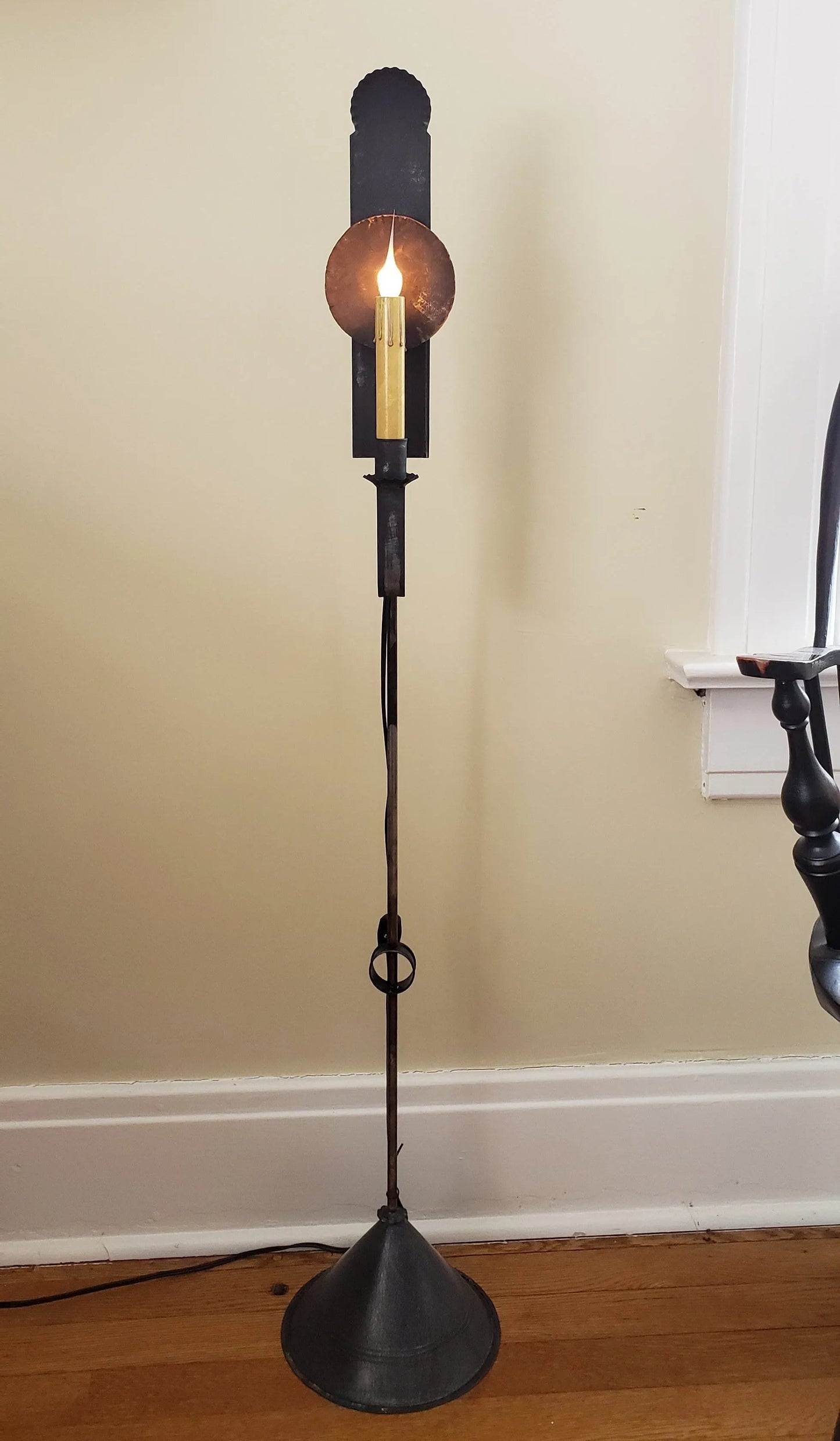 A black mini tin floor lamp with a lit wick, placed on a wooden floor against a wall.