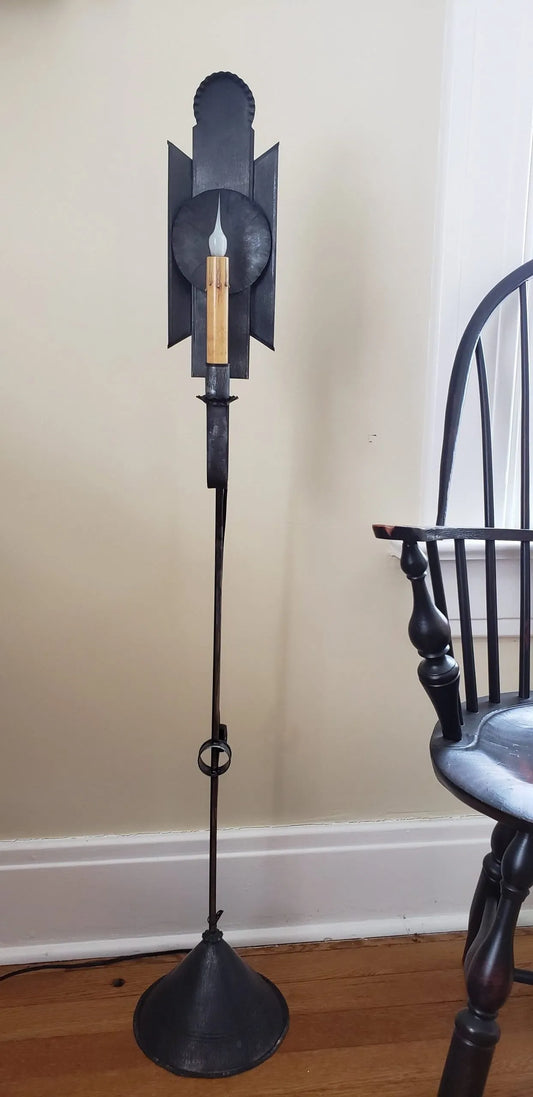 A mini tin floor lamp with a black finish, featuring a triangular base and a vertical post with a lantern-like top. The base is placed on a floor against a wall, with a chair and part of another furniture piece visible to the side.
