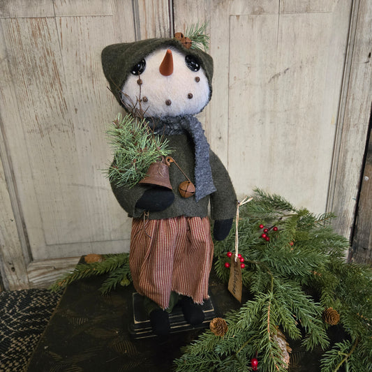 Decorative snowman figure with a green coat and brown boots, standing next to a Christmas tree on a wooden floor.