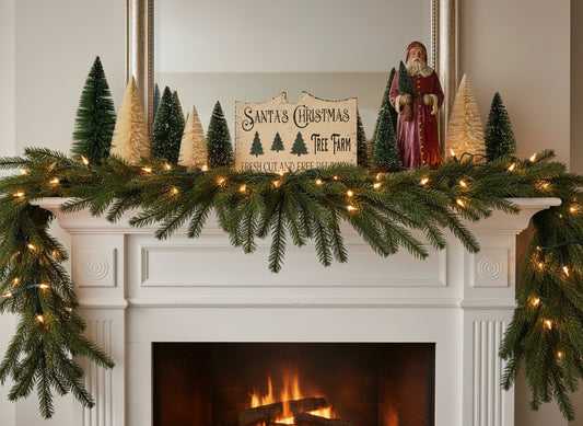 Decorative Christmas mantel with garland, trees, and a sign above a fireplace.