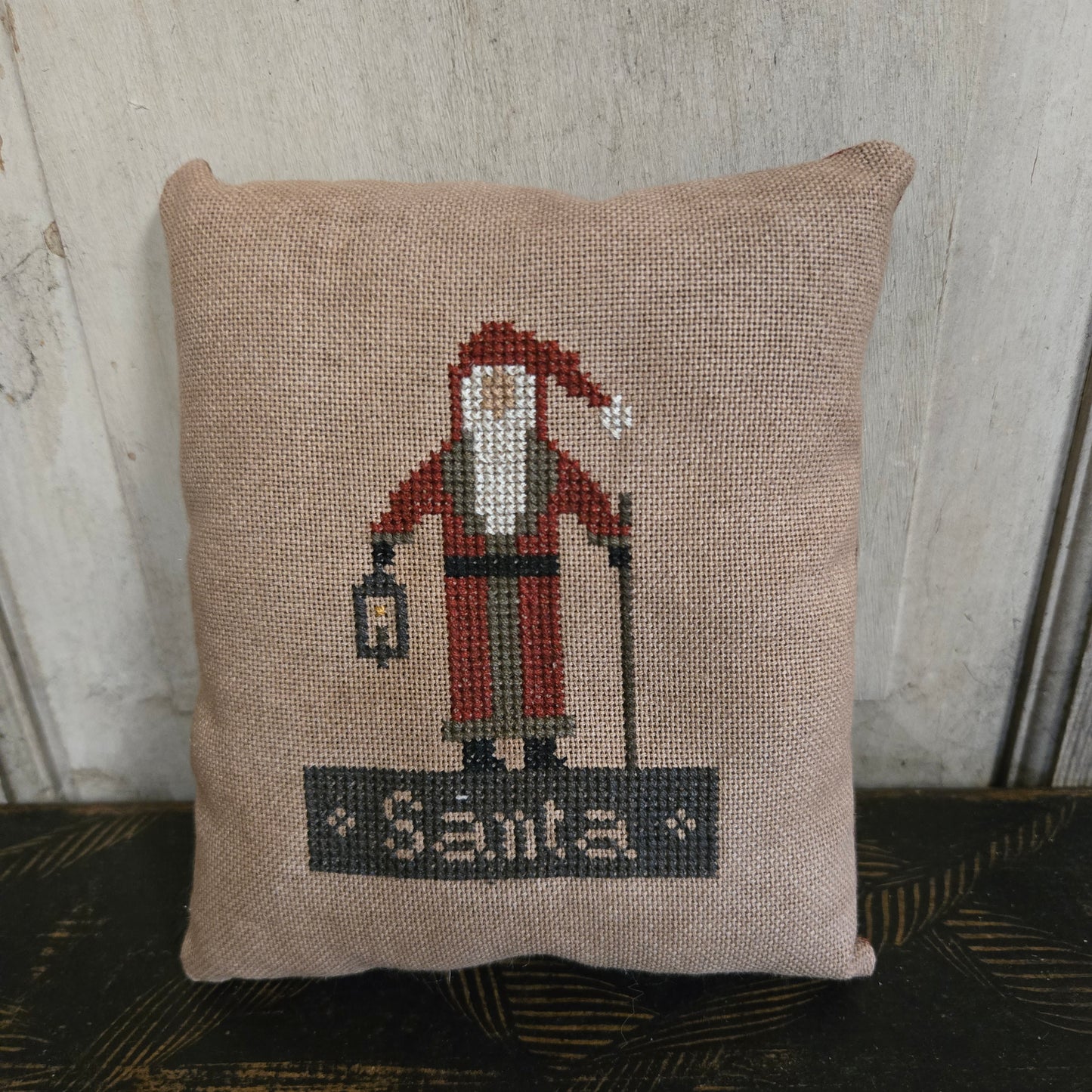 Decorative pillow with a cross-stitched design of a vintage airplane and 'Santas' text, placed against a neutral background.