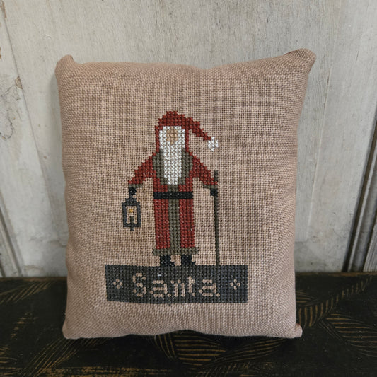 Decorative pillow with a cross-stitched design of a vintage airplane and 'Santas' text, placed against a neutral background.