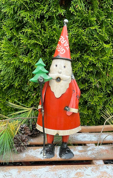 A metal figurine of Santa Claus holding a Christmas tree on a stick, with a red suit and hat, on a wooden surface with greenery in the background.