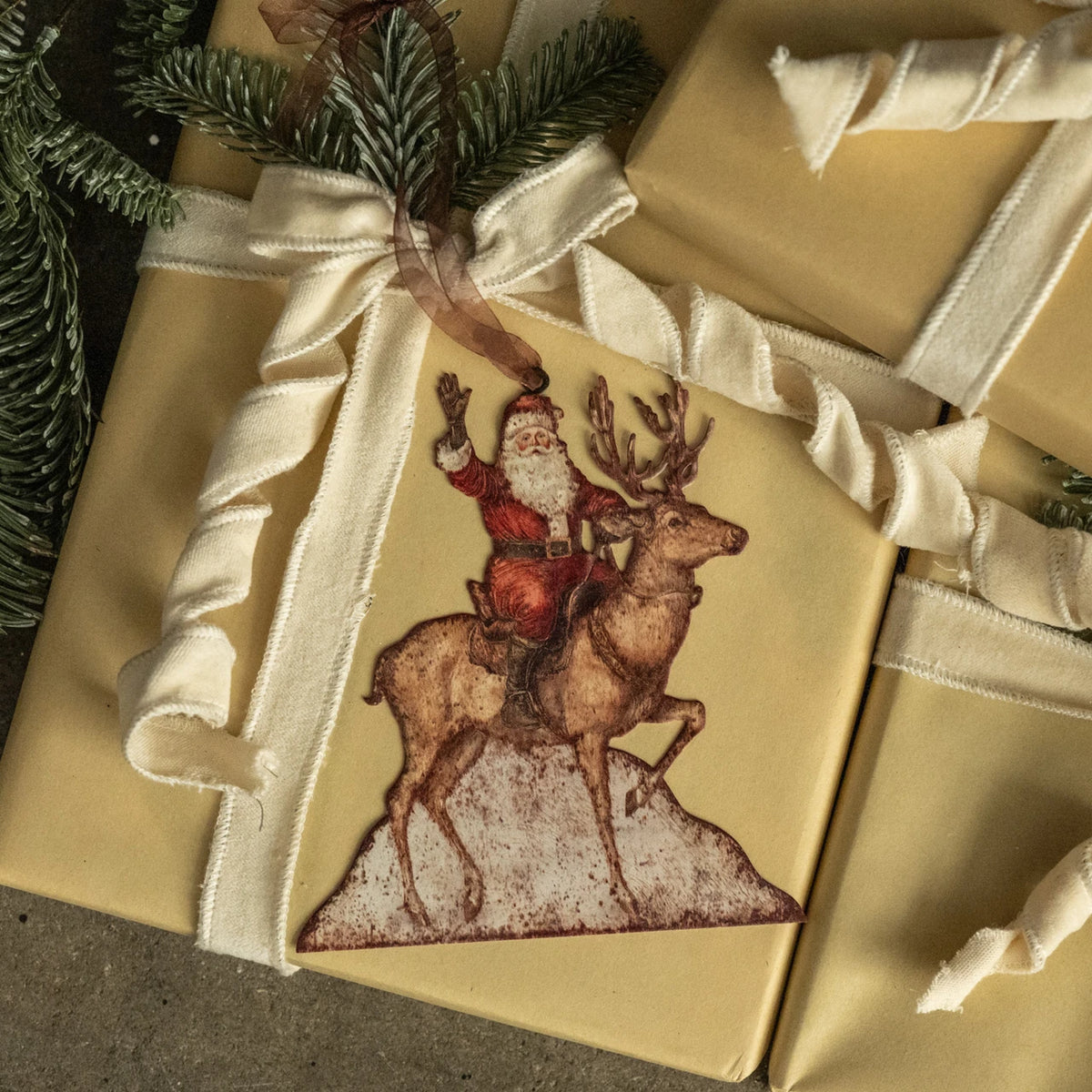 A Christmas-themed gift tag featuring a vintage illustration of Santa Claus riding a reindeer, attached to a kraft paper gift box with a cream ribbon and a pine sprig.