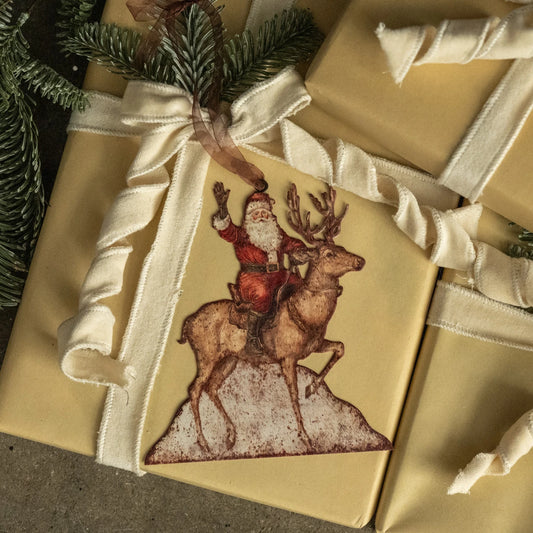 A Christmas-themed gift tag featuring a vintage illustration of Santa Claus riding a reindeer, attached to a kraft paper gift box with a cream ribbon and a pine sprig.