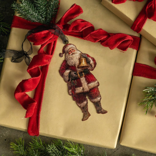 A vintage-style Santa Claus ornament or sticker applied to a gold gift box wrapped with a large red ribbon, surrounded by pine branches.