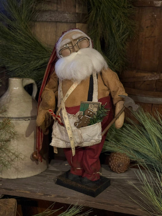Vintage-style Santa Claus figure with a beard, glasses, and a bag on a wooden surface with greenery and a white jug.