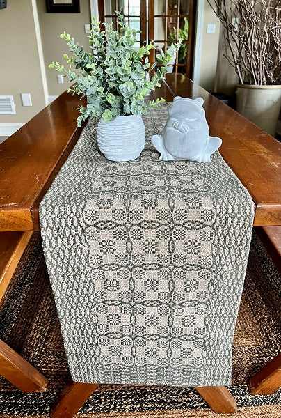 A rectangular green and linen placemat with a geometric pattern, displayed on a wooden dining table with a vase of flowers and a gray fabric object to the side.