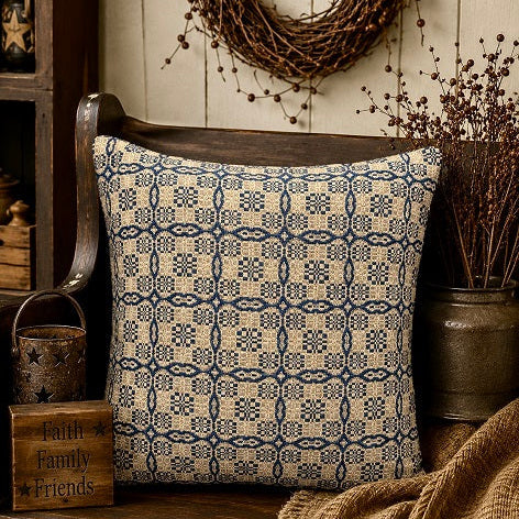 A square throw pillow with a navy blue geometric chain-link pattern on a cream background, styled on a rustic wooden bench in a cozy, country-themed room setting.
