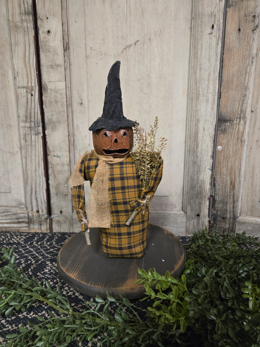 A scarecrow with a paper mache head, featuring a checkered shirt and a burlap sack, displayed on a wooden surface with greenery in the background.