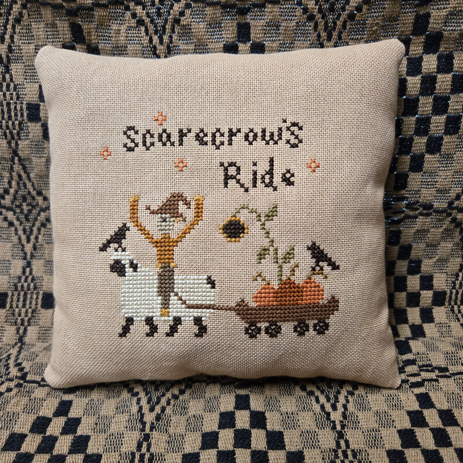A square beige linen throw pillow featuring a cross-stitch design of a scarecrow riding a white sheep to pull a wagon with pumpkins, under the text 'Scarecrow's Ride'. The pillow is displayed on a black and white checkered couch.