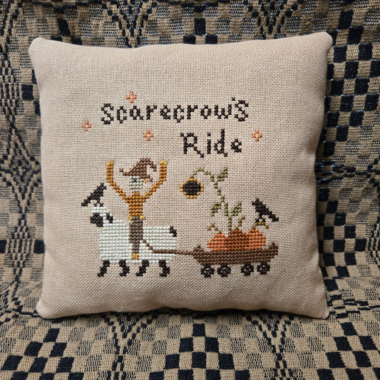 A square beige linen throw pillow featuring a cross-stitch design of a scarecrow riding a white sheep to pull a wagon with pumpkins, under the text 'Scarecrow's Ride'. The pillow is displayed on a black and white checkered couch.