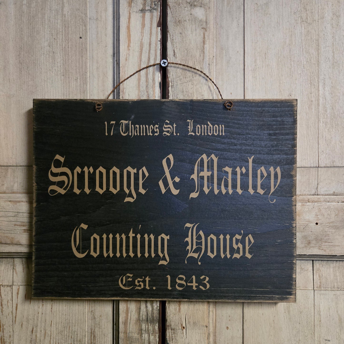 Black sign with wire hanger with lettering that says 'Scooge & Marley Counting House'.