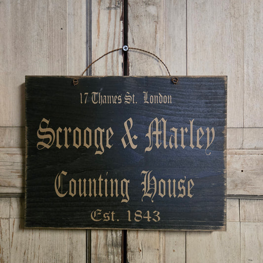 Black sign with wire hanger with lettering that says 'Scooge & Marley Counting House'.