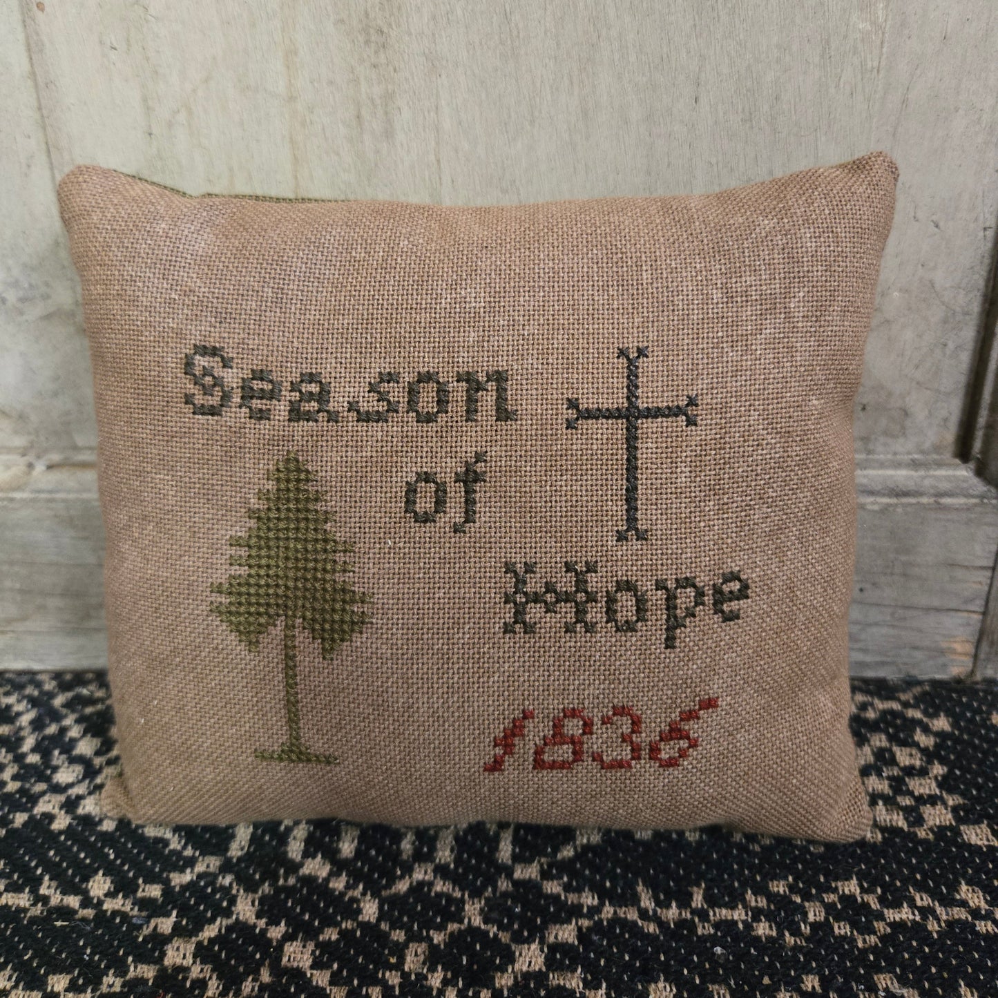 A square cross stitch pillow with the text 'Season of Hope 1836' and a cross and tree motif, displayed against a wooden background.