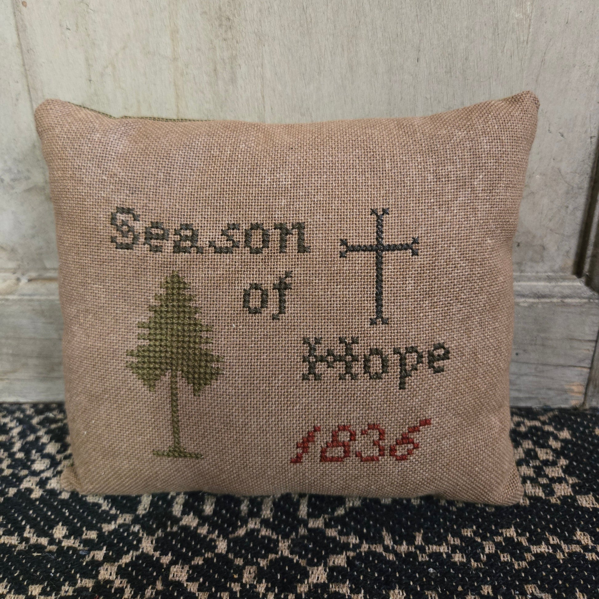 A square cross stitch pillow with the text 'Season of Hope 1836' and a cross and tree motif, displayed against a wooden background.