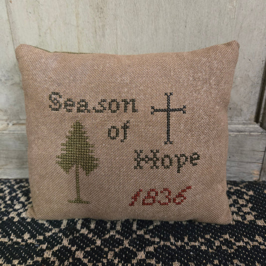 A square cross stitch pillow with the text 'Season of Hope 1836' and a cross and tree motif, displayed against a wooden background.