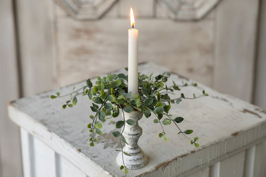 White candle on a wooden stand with greenery on a rustic surface
