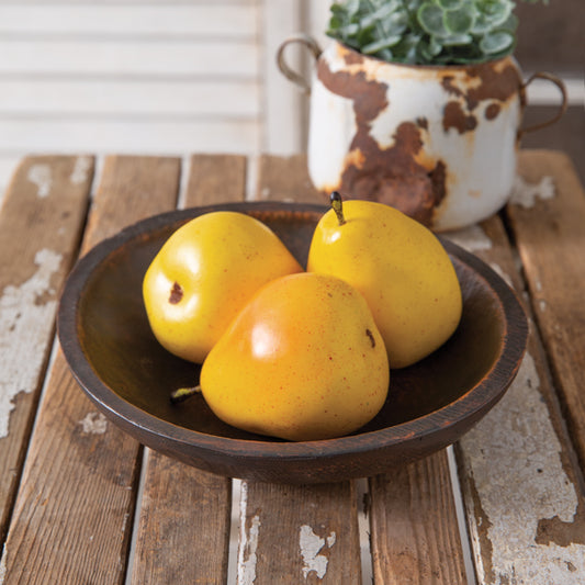 A group of three ripe yellow apples placed in a brown, oblong shallow bowl made of resin, positioned on a wooden surface with a rustic metal object and a potted plant in the background.
