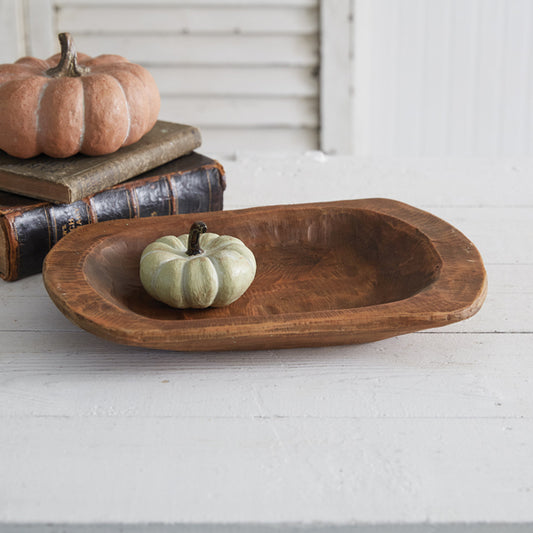 A wooden-look resin shallow dough bowl placed on a white surface, with a pumpkin and a small candle placed in it, next to a book and a pumpkin.