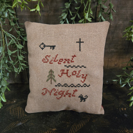 Decorative pillow with cross-stitched designs and text, surrounded by greenery.