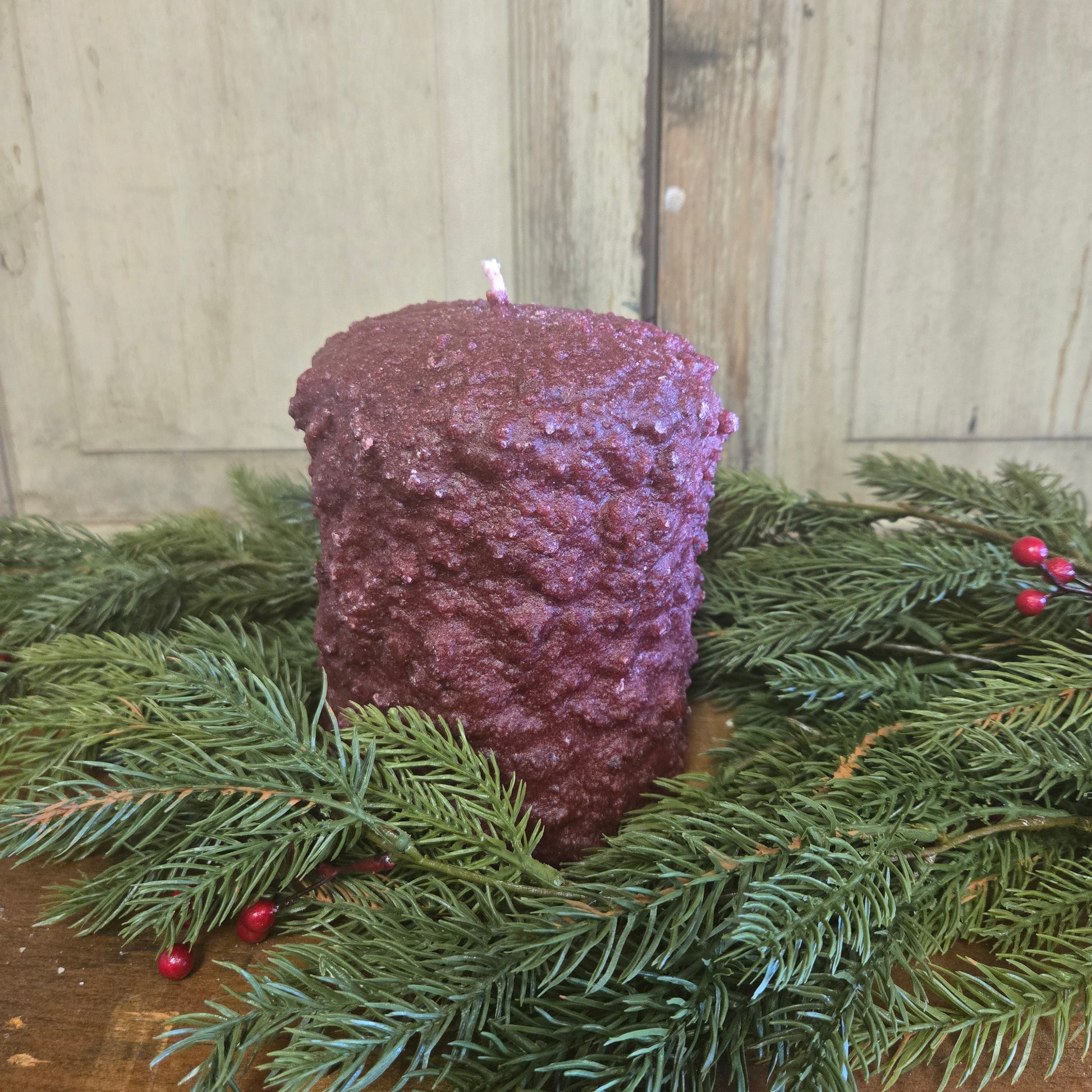 A burgundy colored hearth candle surrounded by greenery against weathered wood