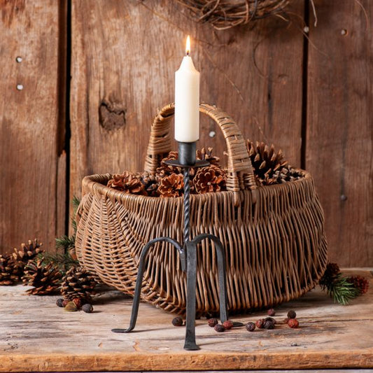 A wrought iron candle holder with a single taper candle, placed on a wicker basket filled with pine cones, set against a rustic wooden backdrop.