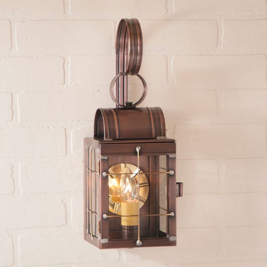 Antique copper wall lantern with a clear glass cover and brass trim, mounted on a white brick wall, emitting a warm light.