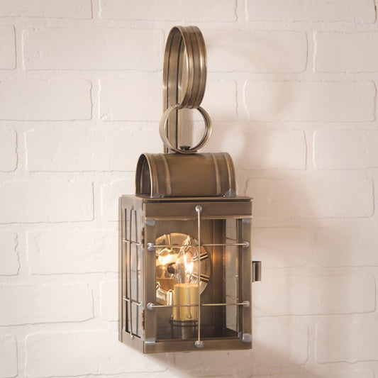A single wall lantern in weathered brass with a clear glass cover and a visible light bulb inside, mounted on a white brick wall.