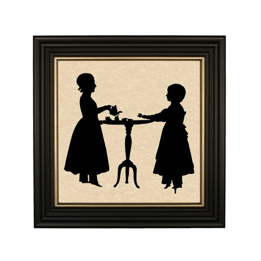 A framed paper cut silhouette of two figures, resembling sisters, having a tea party. The silhouette is black with gold trim, set against a beige background, enclosed in a black wood frame.