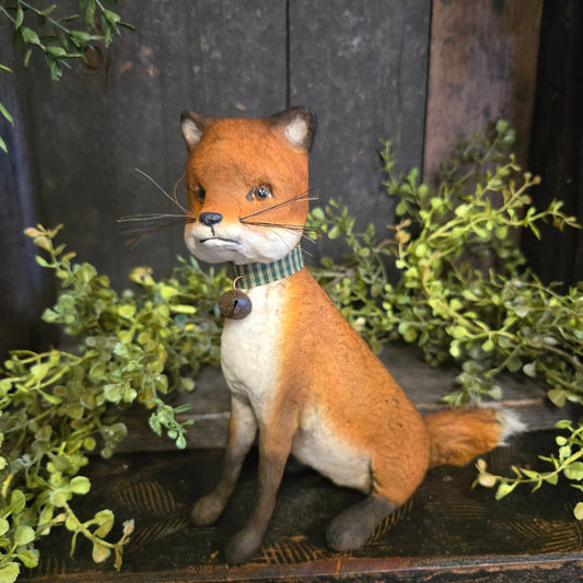 Paper mache fox with a striped tie among green plants on a wooden surface