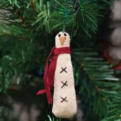 A plush, beige snowman ornament with stitched features, wearing a red felt scarf, hanging from a green pine tree.