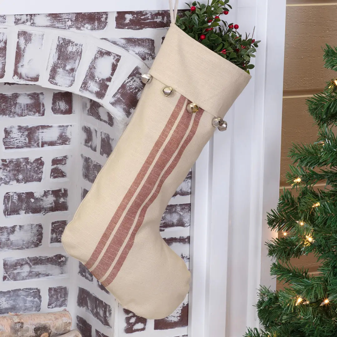A beige linen Christmas stocking with two vertical dusty rose stripes and silver jingle bells at the top, hanging in front of a white brick fireplace next to a decorated Christmas tree.
