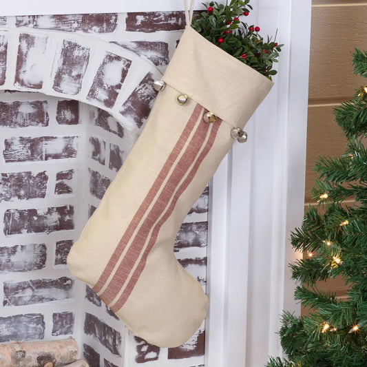 A beige linen Christmas stocking with two vertical dusty rose stripes and silver jingle bells at the top, hanging in front of a white brick fireplace next to a decorated Christmas tree.