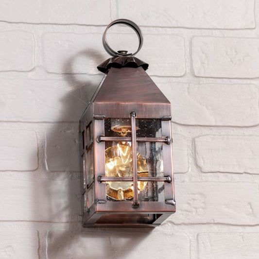 A small outdoor wall light fixture with a solid antique copper finish, featuring a glass door and a blackened copper frame, mounted on a white brick wall.