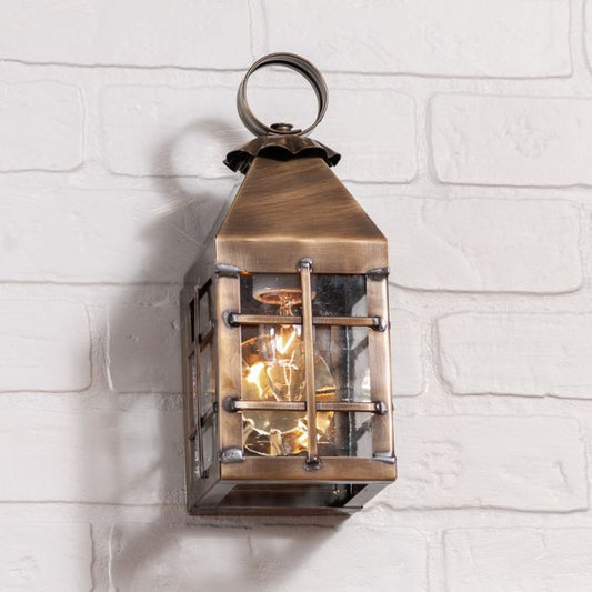 A small outdoor wall light fixture in solid weathered brass, hanging on a white brick wall, with a visible lit light inside.
