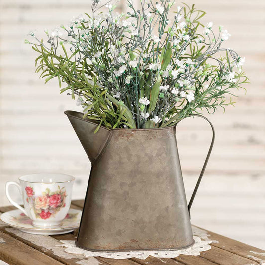 Metal pitcher with artificial flowers on a table next to a teacup and saucer
