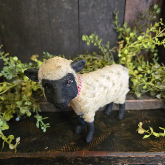 Plush sheep toy with black face and white wool, hanging from a wooden frame with greenery.
