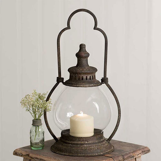 Glass globe lantern in iron holder with a tealight on a wooden table with a jar of flowers next to it
