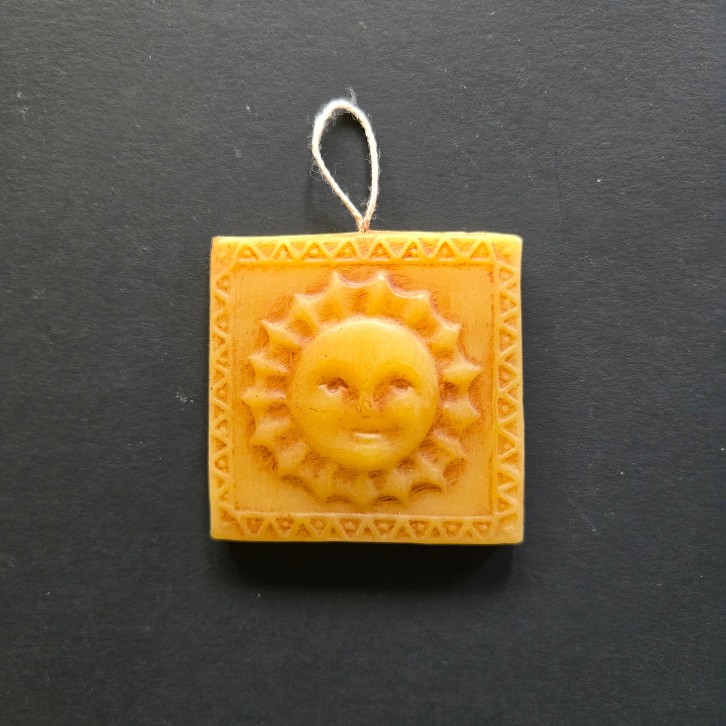 Square beeswax ornament with sun design and string on a black background