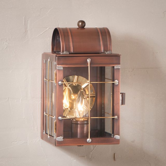 A small wall-mounted lantern in antique copper color with a clear glass front and a lit candle inside, creating a warm glow.